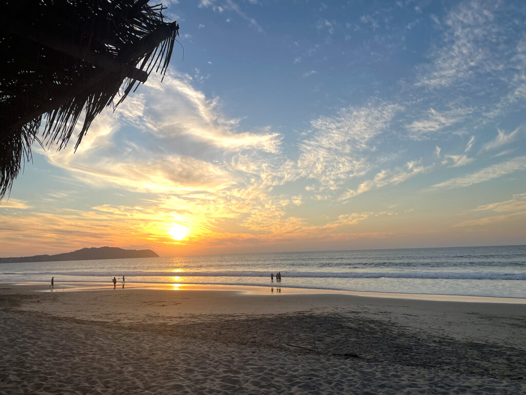 A sunset on a beach, with a blue and orange sky, with people in the distance walking and a palm roof just visible in the side of the photo